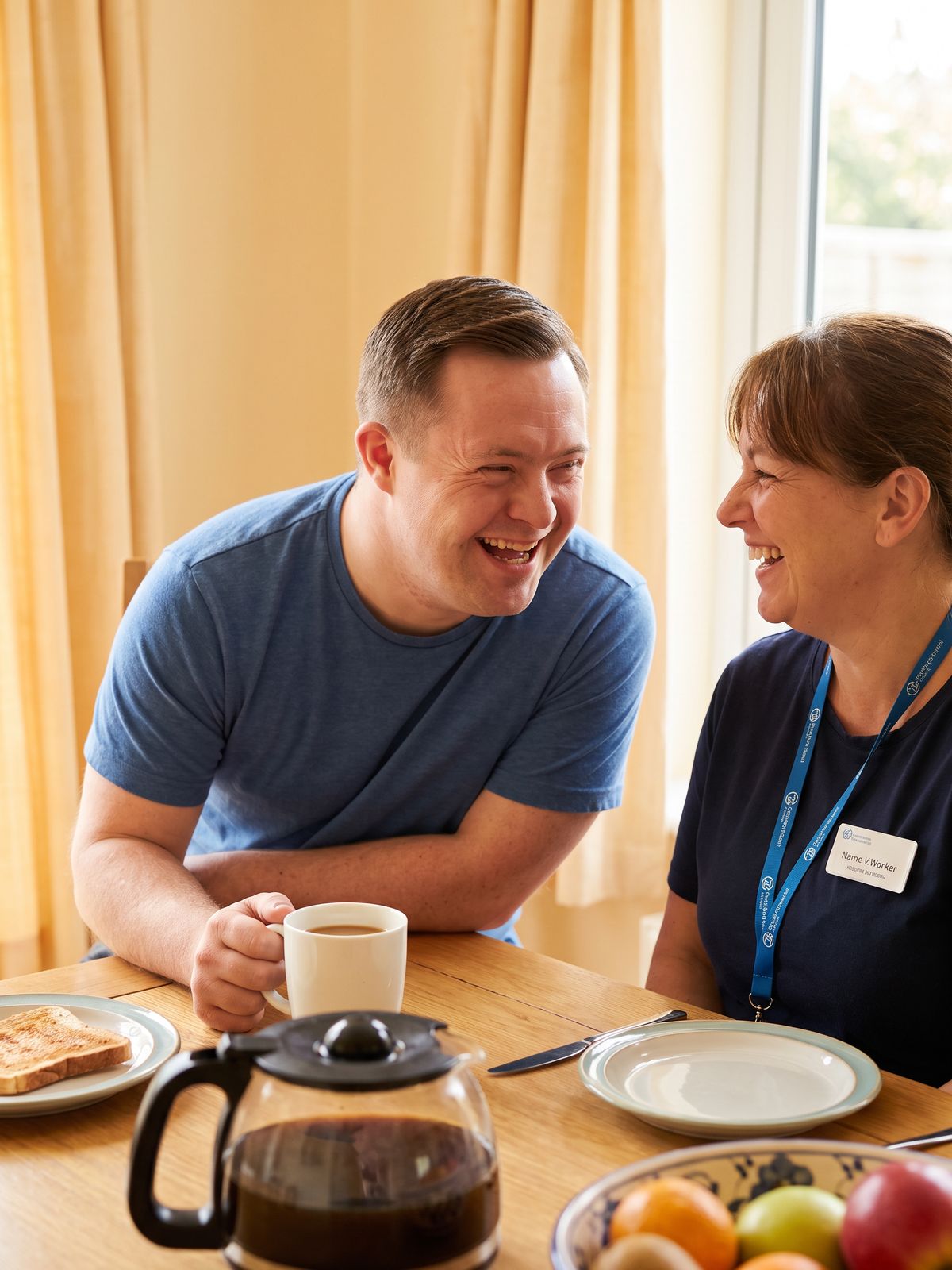 A man with Down syndrome in a blue t-shirt laughs over coffee and toast with his support worker, who wears a lanyard name badge, at a sunny table