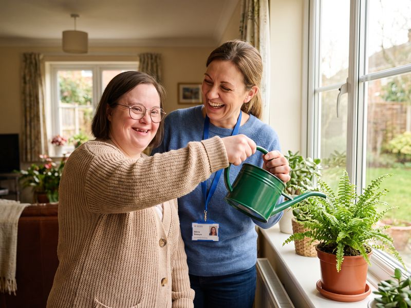 A woman with Down syndrome in glasses and a cardigan smiles as she waters houseplants by a sunny window with her caregiver helping