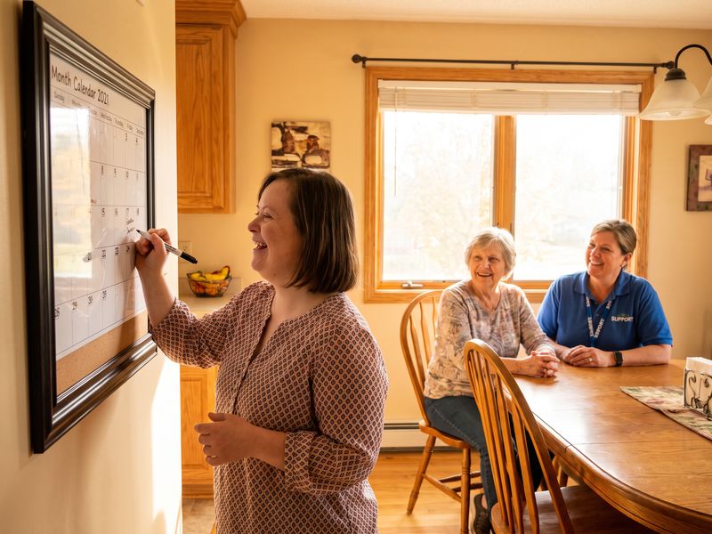 A young woman with Down syndrome writes her weekly plan on a wall calendar while her mother and a support worker smile from the table